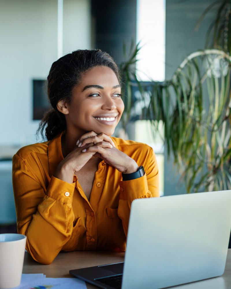 Portrait of dreamy african american businesswoman sitting at desk in office and thinking while working on laptop, looking away with pleased face expression, copy space